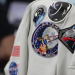 A gray colored jacket with five Artemis mission patches and a NASA insignia hangs on a chair.