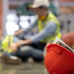 Close Up on Red Hard Hat with Blurred Injured Factory Workers as